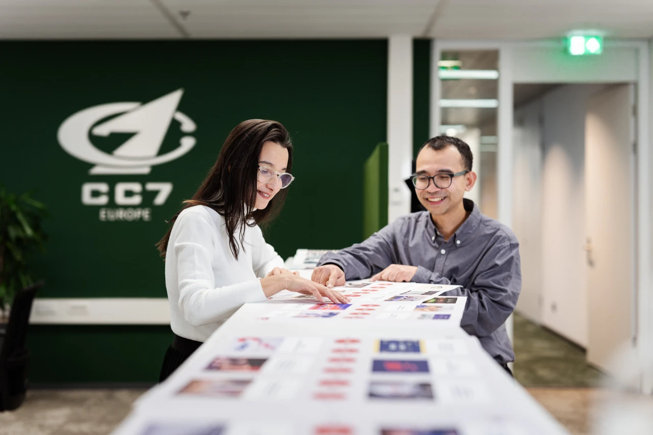 1 woman and 1 man standing checking a sheet with the CC7 Europe logo in the background.
