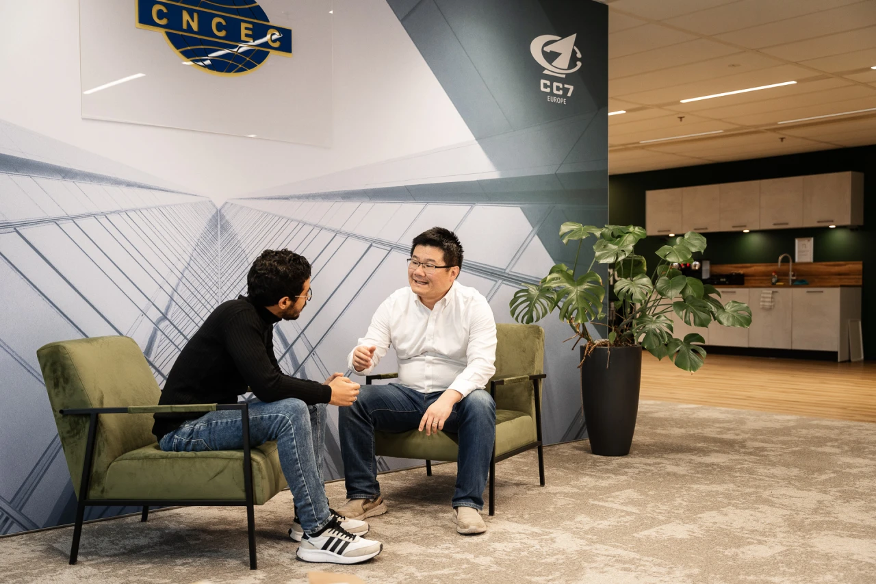 2 men conversing sitting on 2 green armchairs next to a plant 2 men conversing sitting on 2 green armchairs next to a plant