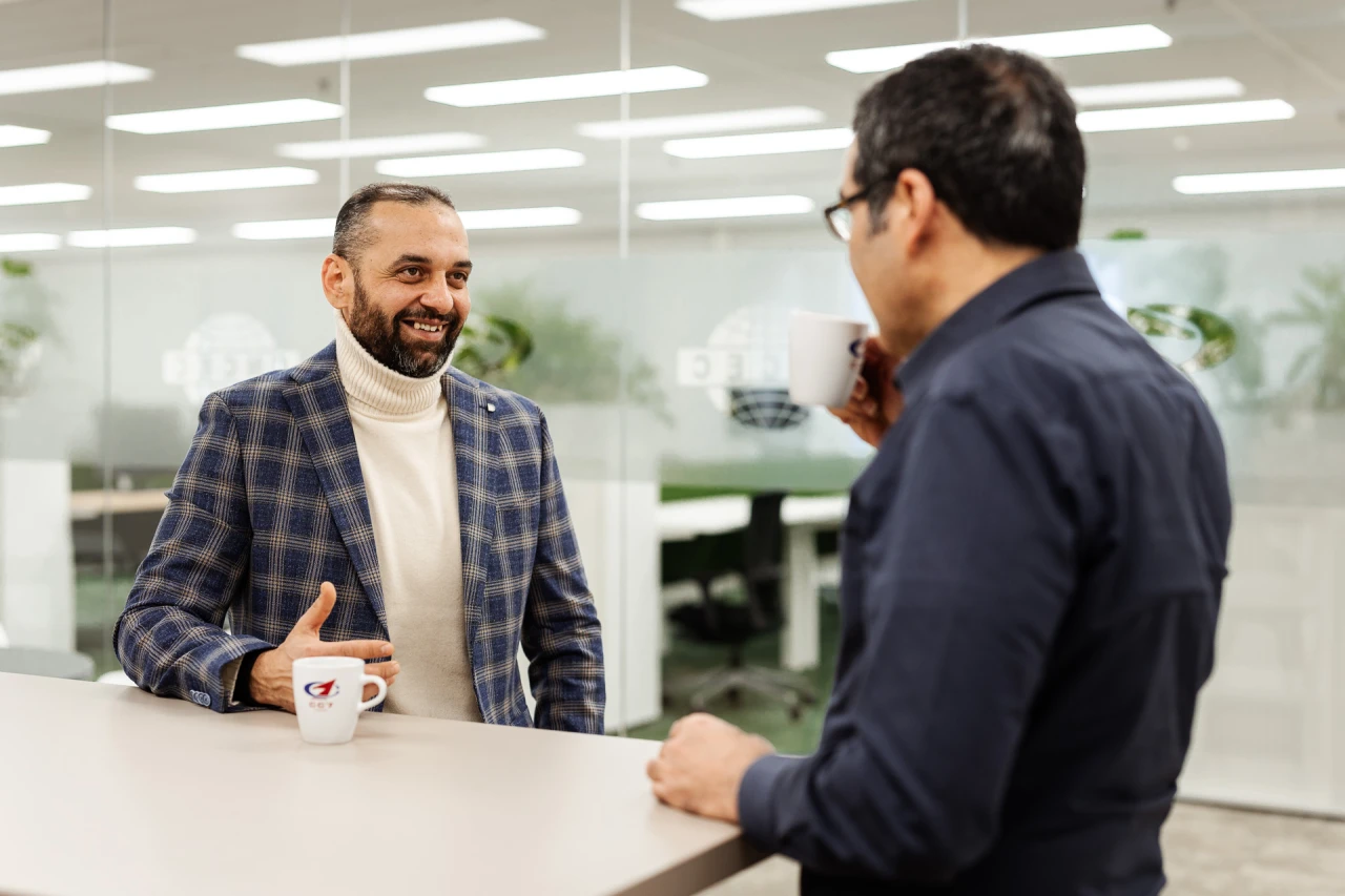 2 smiling men standing and talking while they drink from 2 cups.