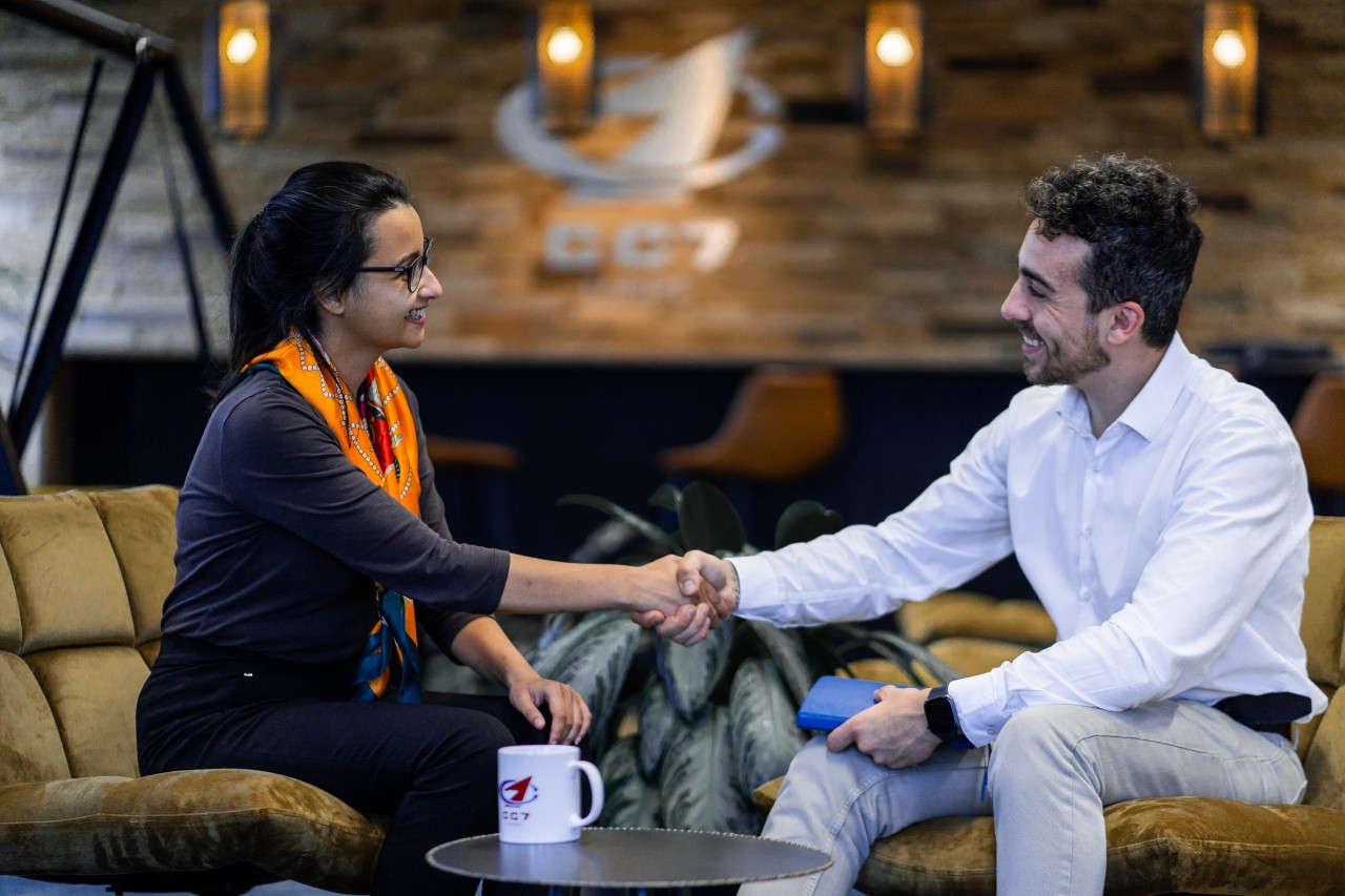 2 smiley peolple shaking hands while sitting on 2 chairs with a branded cup on the small table.