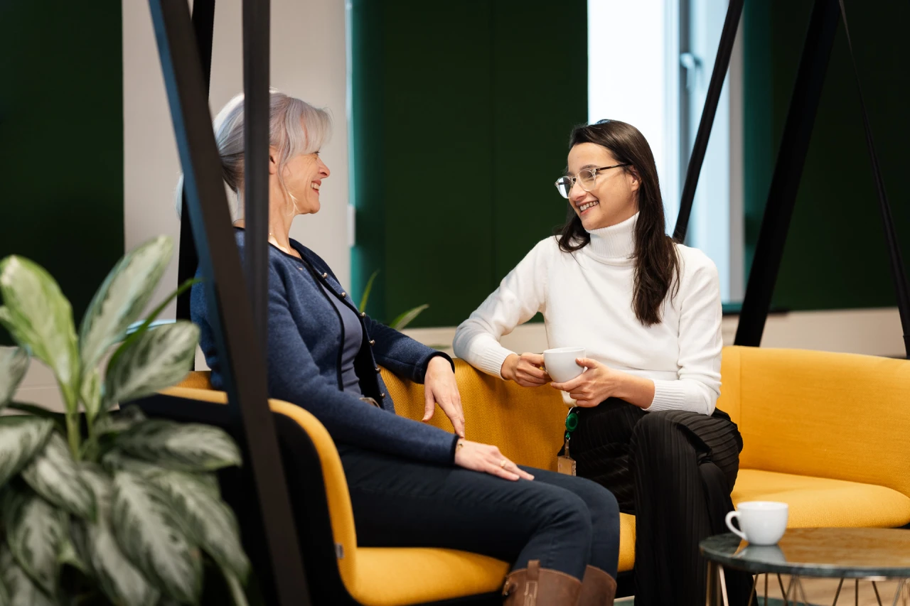 2 smiling ladies conversing and smiling on a yellow swing holding 1 cup of coffee.