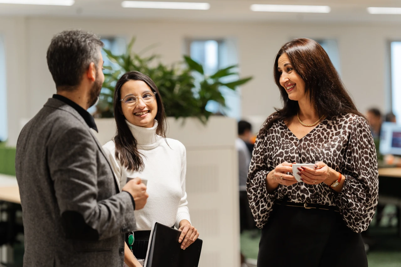 3 people standing and smile while conversing in office and holding 1 agenda and 1 cup.