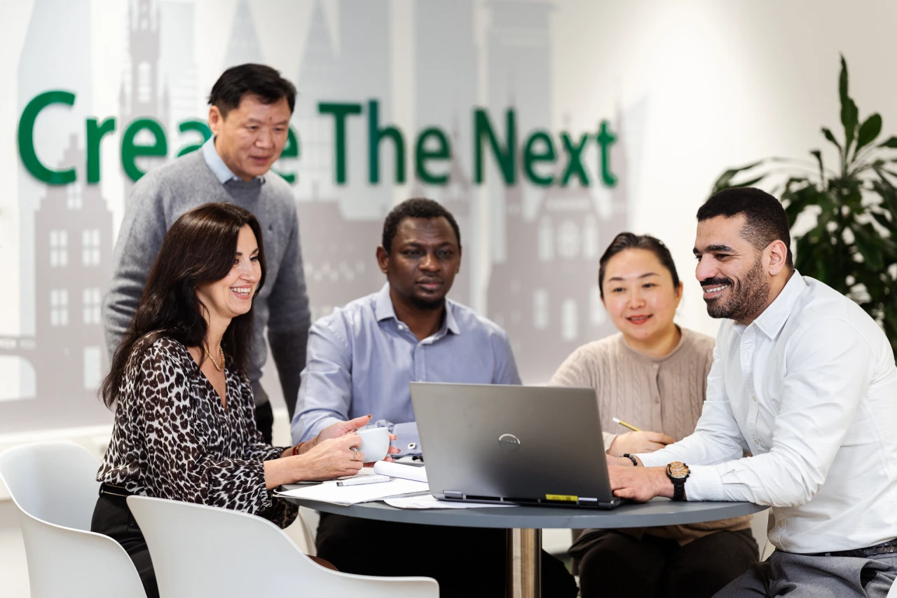 5 people aound a round table checking a laptop and conversing with each other.