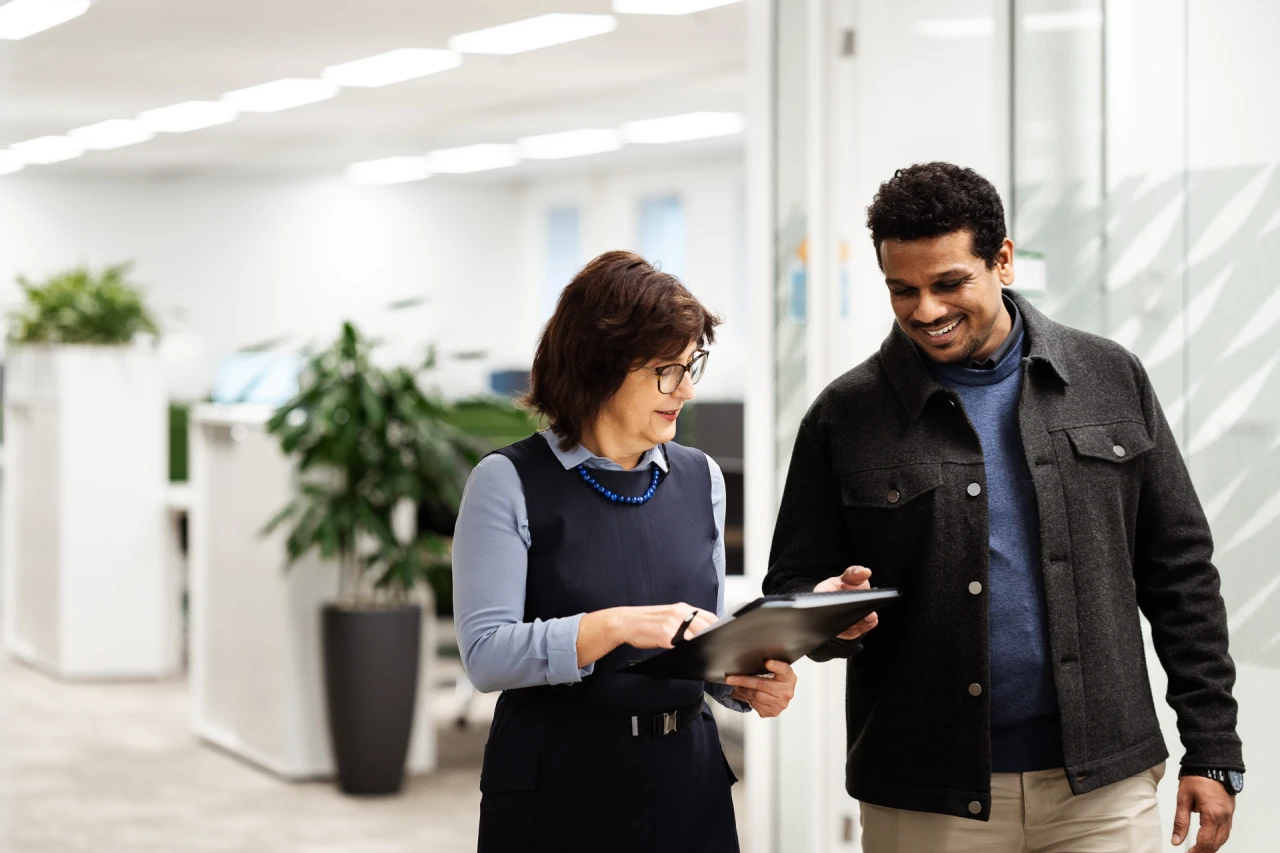 1 women and 1 man walking in office and conversing with each other while they check a document.